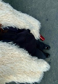 Casaco de pelo sintético, macio e branco, com uma textura longa e ondulada, exibido sobre um hoodie preto e calças. Sapatos pretos com um acabamento brilhante visíveis em baixo.