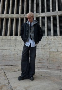 Jeune personne aux cheveux blonds portant une veste noire, un sweat à capuche gris, une chemise blanche, une cravate noire, un pantalon large noir et des chaussures à plateforme noires, debout en plein air.