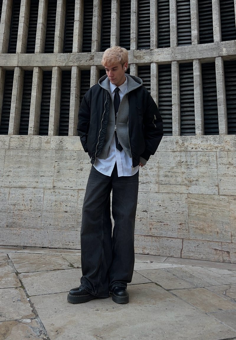 Jeune personne aux cheveux blonds portant une veste noire, un sweat à capuche gris, une chemise blanche, une cravate noire, un pantalon large noir et des chaussures à plateforme noires, debout en plein air.