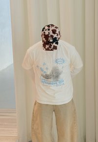 Personne portant un pantalon beige, un t-shirt blanc style vintage avec un graphisme Ibiza, et une casquette New York Yankees à motif vache, debout les mains derrière le dos.