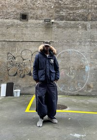 Námořnická parka s kapucí lemovanou kožešinou, spárovaná s kalhotami širokého střihu s bílými bočními pruhy. Černobílé kostkované boty. Pozadí s graffiti.