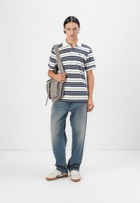 Polo a righe bianco, blu navy e verde; abbinato a jeans dritti azzurri e sneakers bianche; porta una borsa a tracolla grigia.