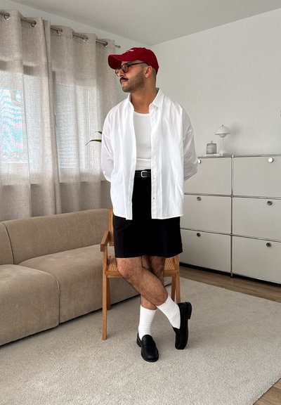 Camisa blanca de botones sobre una camiseta blanca sin mangas, falda negra, mocasines negros y gorra roja. El fondo presenta un sofá beige y mobiliario moderno.