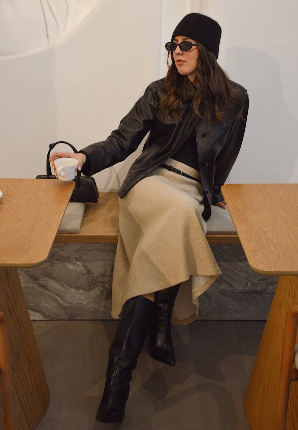 Veste en cuir noire, haut noir ajusté, jupe asymétrique beige, bottes noires montant jusqu'aux genoux, lunettes de soleil rondes et sac à main noir.