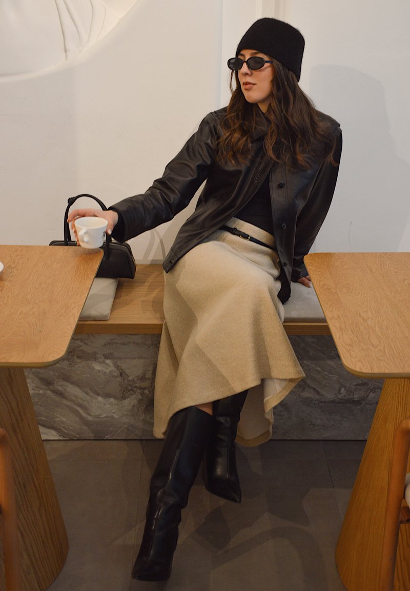 Veste en cuir noire, haut noir ajusté, jupe asymétrique beige, bottes noires montant jusqu'aux genoux, lunettes de soleil rondes et sac à main noir.