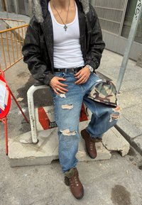 Črna jakna iz umetnega usnja z odebeljenim krznenim kapuco, bela majica brez rokavov, obrabljene modre kavbojke in rjavi pohodni škornji. Kamuflažna kapa počiva na kolenu.