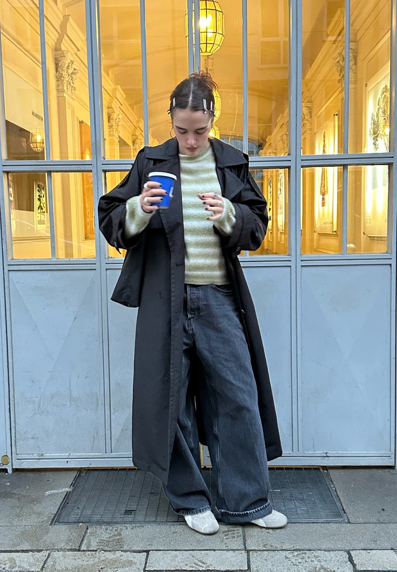 Trench nero sopra un maglione verde chiaro a righe, abbinato a jeans scuri a gamba larga e scarpe slip-on grigie. Tiene in mano una tazza da caffè blu.