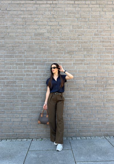 Mujer con gafas de sol, camisa negra, pantalones anchos marrones, zapatillas blancas, sosteniendo un bolso, de pie junto a una pared de ladrillos grises en una acera.