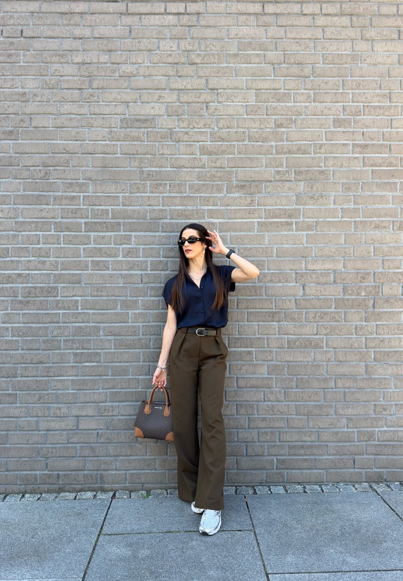 Frau mit Sonnenbrille, schwarzem Hemd, brauner weiter Hose, weißen Sneakers, hält eine Handtasche, steht vor einer grauen Ziegelmauer auf dem Bürgersteig.