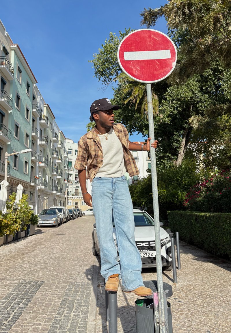 Eine Person steht auf einem Pfahl neben einem Zutritt verboten-Schild, trägt ein karriertes Hemd, ein weißes Hemd, hellblaue Jeans und braune Stiefel in einem städtischen Umfeld.