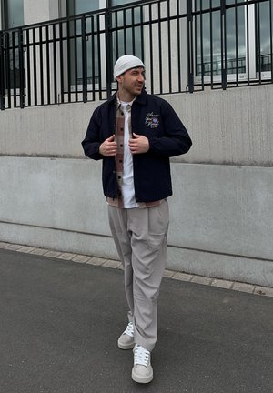 Joven con pantalones beige, chaqueta de varias capas, gorro blanco y zapatillas, de pie en una acera junto a una pared de hormigón con barandillas de metal.
