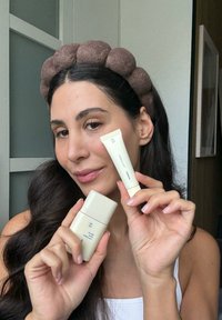Due tubetti per la cura della pelle color beige chiaro sono tenuti in mano, con testo minimalista. Un morbido e rotondo cerchietto marrone adorna dei capelli scuri e mossi.