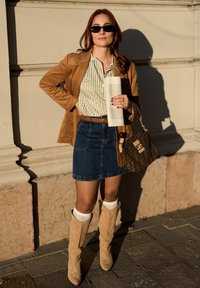 Καφέ σουέτ σακάκι, ριγέ μπλούζα, τζιν φούστα, μπεζ μπότες με λεπτομέρειες από κορδόνια, τυπωμένη τσάντα και κρατώντας ένα τυλιγμένο αντικείμενο.