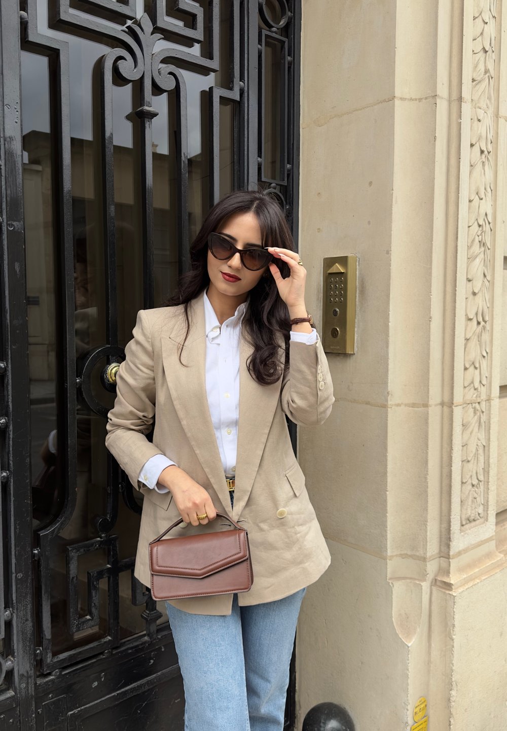 Blazer beige, chemise blanche à boutons, jeans bleus, tenant un sac à main géométrique marron. Lunettes de soleil noires, porte texturée avec des détails ornés.