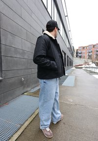 Homme portant une veste noire avec un col en fourrure, un jean ample bleu clair et des baskets, debout sur un trottoir à côté d'un bâtiment moderne gris.