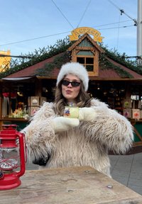 Une personne vêtue d'un manteau crème duveteux et de gants blancs tient une tasse jaune décorée, à côté d'une lanterne rouge, sur fond de marché festif.