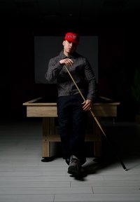 Un jeune homme portant une casquette rouge et une veste à motifs s'appuie sur une queue de billard, debout devant une table de billard en bois dans une pièce faiblement éclairée.