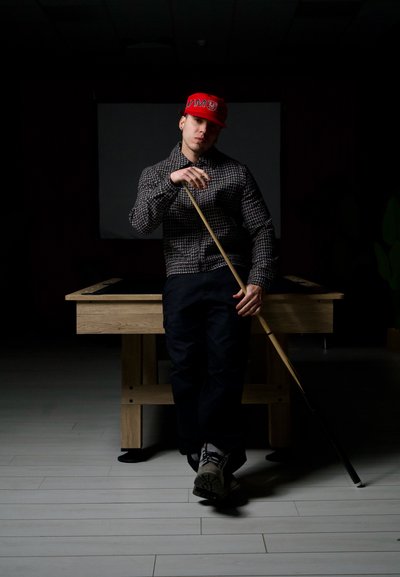 Joven con gorra roja y chaqueta estampada apoyado en un taco de billar, de pie frente a una mesa de billar de madera en una habitación con poca luz.