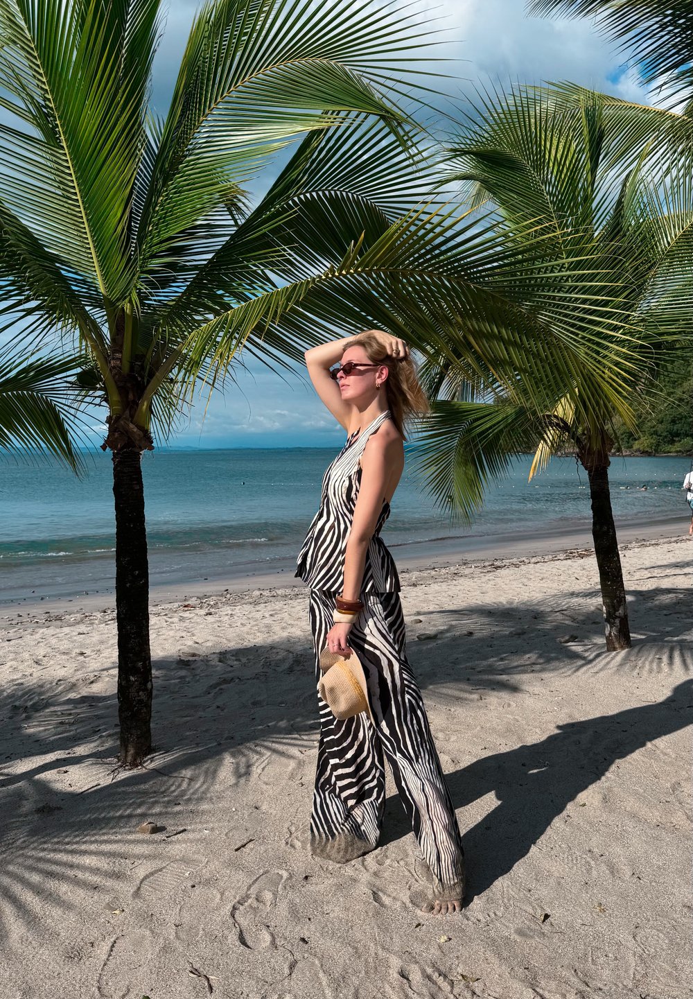 Débardeur rayé zébré et pantalon large noirs et blancs. Chapeau de paille beige clair tenu à la main. Plage de sable avec palmiers et océan en arrière-plan.