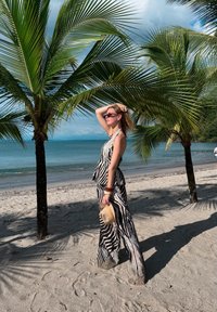 Débardeur rayé zébré et pantalon large noirs et blancs. Chapeau de paille beige clair tenu à la main. Plage de sable avec palmiers et océan en arrière-plan.