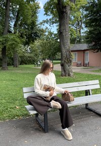 Beige cardigan, bruine wijde broek, crèmekleurige instap schoenen met witte accenten. Houdt een modetijdschrift vast, zittend op een houten bank in een park.