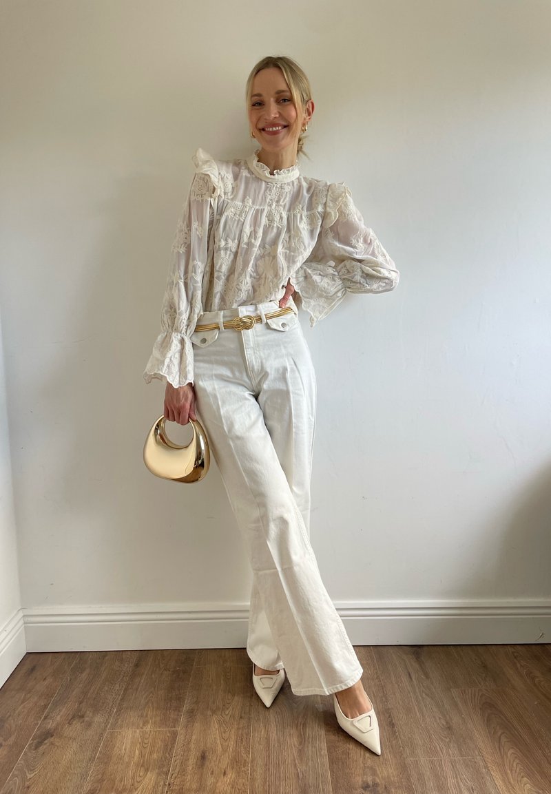 Femme debout contre un mur blanc, portant un chemisier en dentelle blanche transparente, un pantalon blanc taille haute, des chaussures blanches pointues et tenant un petit sac à main doré.