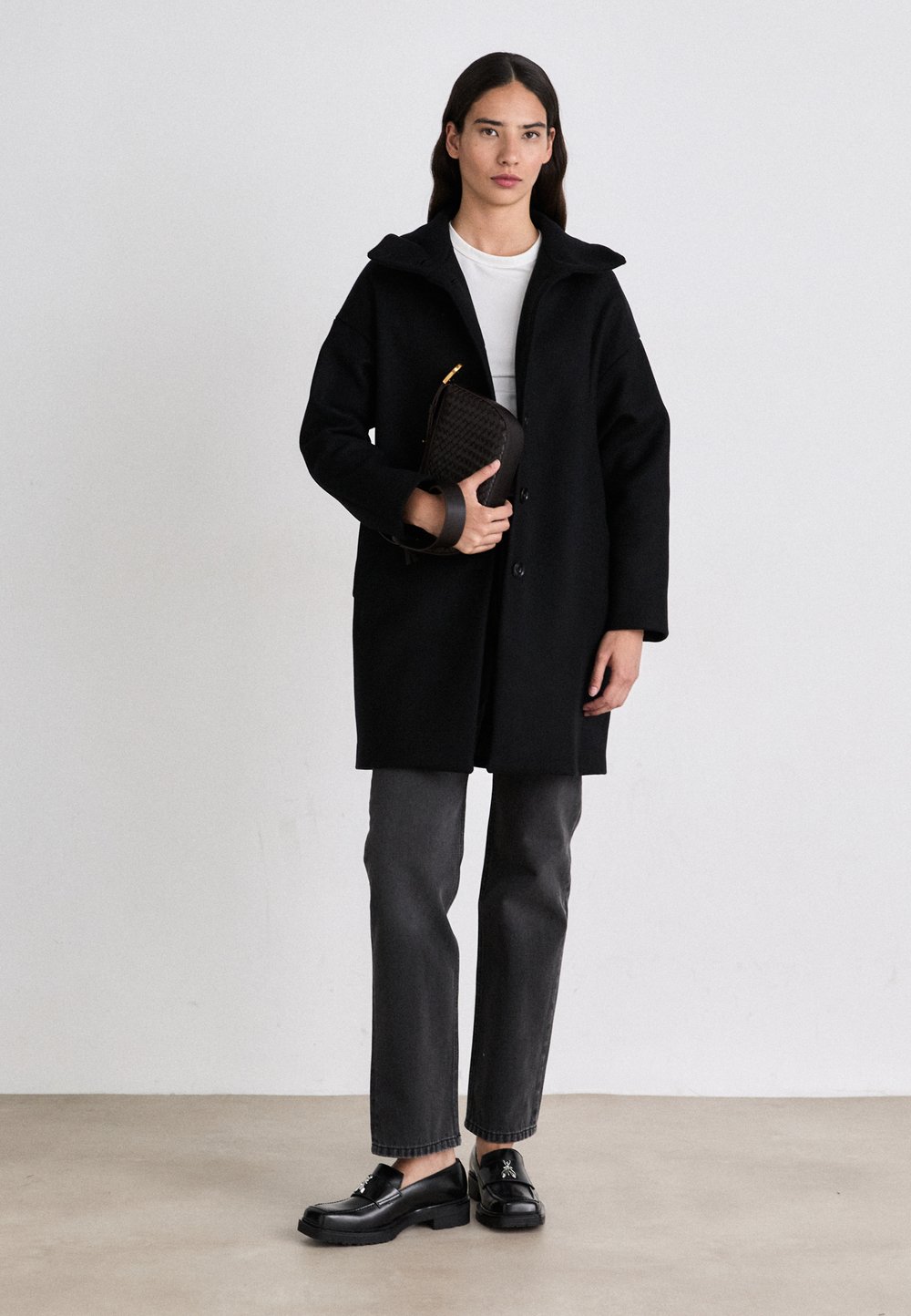 Zwarte wollen jas met capuchon, knoopsluiting, gedragen over een wit shirt. Gecombineerd met grijze broeken en glanzende zwarte loafers; houdt een geweven clutch vast.