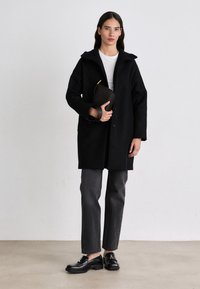 Zwarte wollen jas met capuchon, knoopsluiting, gedragen over een wit shirt. Gecombineerd met grijze broeken en glanzende zwarte loafers; houdt een geweven clutch vast.