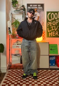 Blouson aviateur noir, débardeur noir, jean ample bleu clair et baskets noires avec des accents néon verts. Accessoires comprenant des colliers en chaîne et des lunettes de soleil.