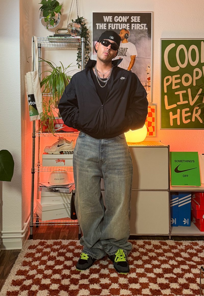 Blouson aviateur noir, débardeur noir, jean baggy bleu clair et baskets noires avec des accents vert néon. Les accessoires incluent des colliers en chaîne et des lunettes de soleil.