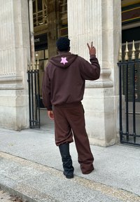 Brun hoodie med en pink stjerne på ryggen, parret med matchende brune sweatpants og sorte knæhøje støvler, sat mod en stenmur.