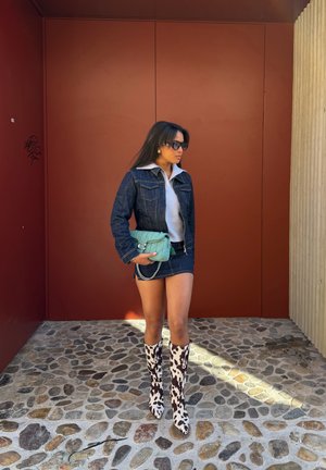 Mujer con chaqueta y falda de mezclilla, top blanco, botas hasta la rodilla con estampado de vaca, gafas de sol y sosteniendo un bolso turquesa, de pie sobre suelo de adoquines.