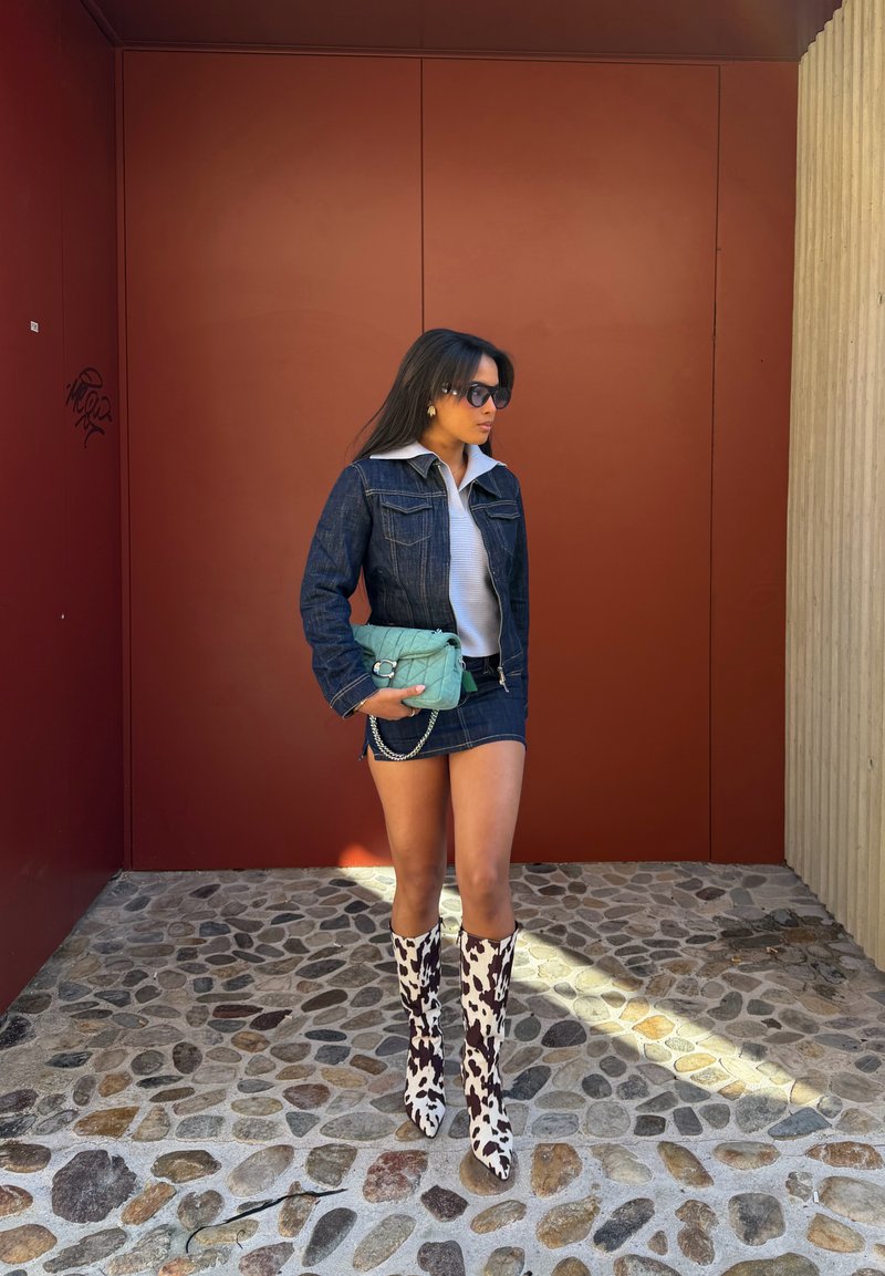 Femme portant une veste en denim et une jupe, un haut blanc, des bottes hautes imprimé vache, des lunettes de soleil, et tenant un sac turquoise, debout sur un sol pavé.