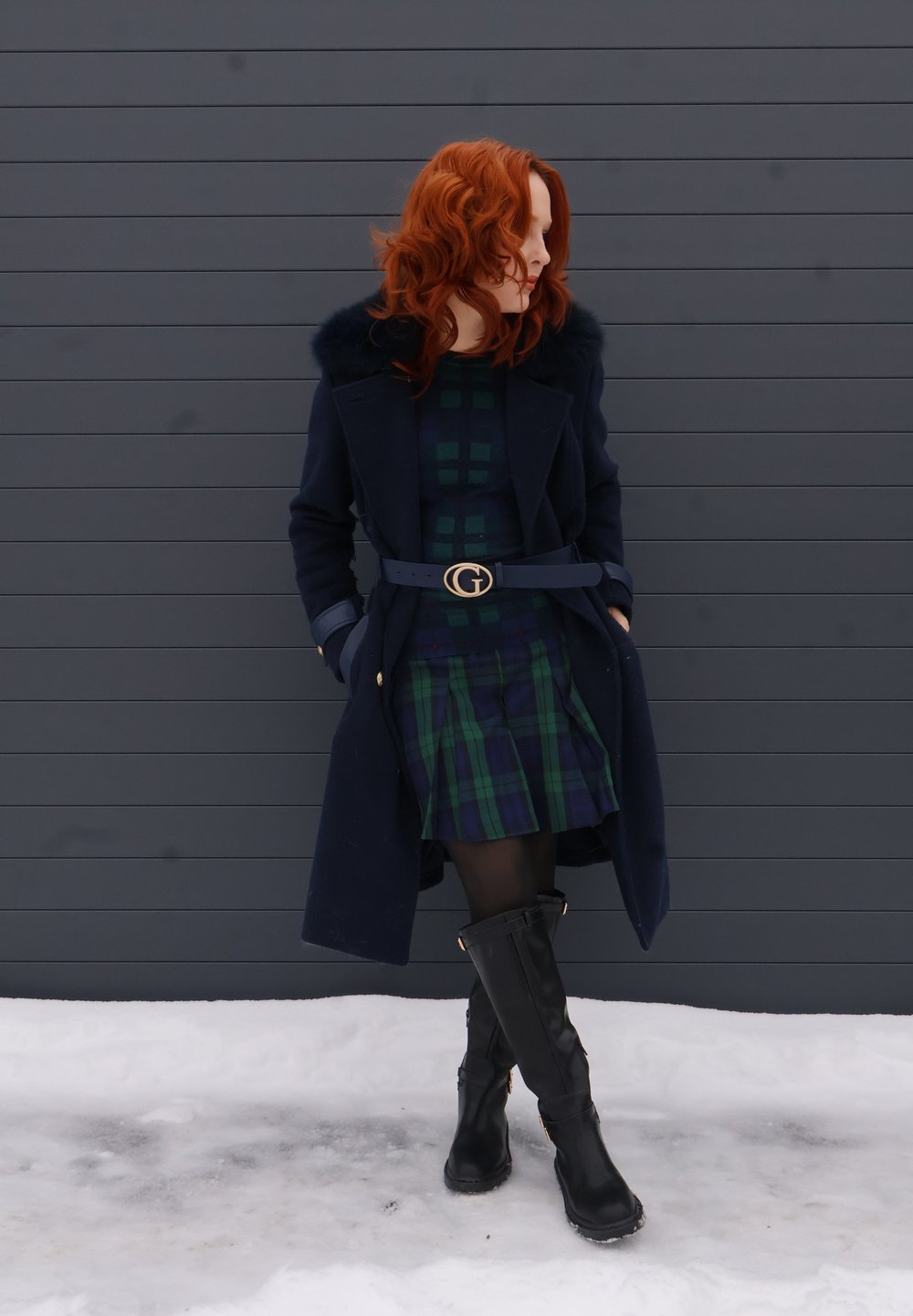 Femme aux cheveux roux portant un manteau bleu marine avec un col en fourrure, une robe à carreaux, des collants noirs et des bottes, debout dans la neige devant un mur gris foncé.