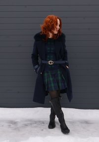 Femme aux cheveux roux portant un manteau bleu marine avec un col en fourrure, une robe à carreaux, des collants noirs et des bottes, debout dans la neige devant un mur gris foncé.