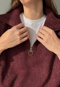 Personne portant une chemise blanche et un pull en tricot bordeaux, tenant le col près d'une fermeture éclair en laiton partiellement zippée.