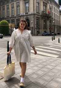 Λευκό βαμβακερό φόρεμα με λεπτομέρειες από τρύπες και φουσκωτά μανίκια; κρατώντας μια διαφανή τσάντα; μπεζ παντόφλες; περπατώντας σε ένα μοτίβο πεζοδρόμιο.