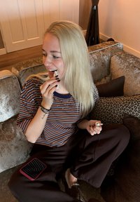 Une femme aux longs cheveux blonds tient un petit tube blanc en  étant assise sur un canapé texturé et à motifs, vêtue d'une chemise à rayures et d'un pantalon marron.