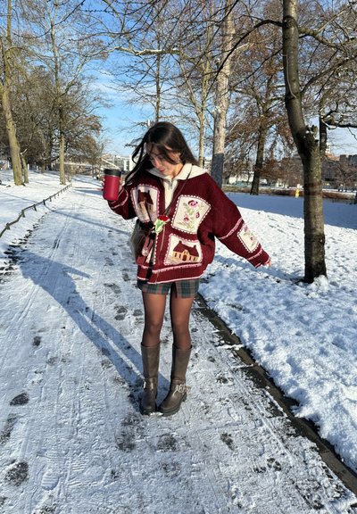 Mujer con suéter burdeos estampado y botas, sosteniendo una taza roja, caminando por un camino cubierto de nieve bordeado de árboles sin hojas en invierno.