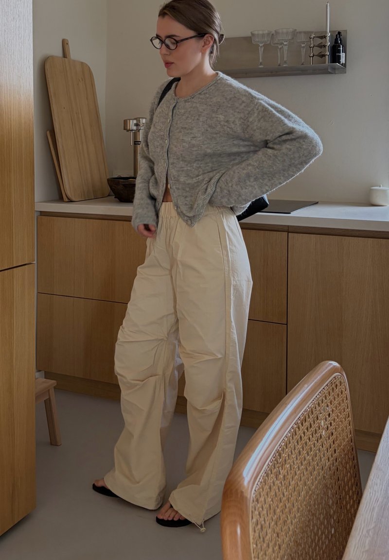 Mujer con gafas, cárdigan gris y pantalones anchos beige, de pie en una cocina de madera con una tabla de cortar y cristalería cerca.