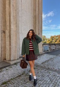 Vrouw in groene jacket, gestreepte shirt, bruine rok, witte sokken en zwarte instappers staat op de kasseien nabij een hoge stenen muur met graffiti.
