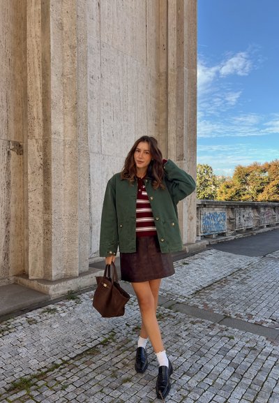 Mujer con chaqueta verde, camisa a rayas, falda marrón, calcetines blancos y mocasines negros se encuentra en un pavimento de adoquines cerca de una alta pared de piedra con graffiti.
