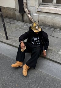 Persona melnas drēbēs un bēšās zābakos, kas sēž uz trotuāra pie ēkas sienas, turot plastmasas krūzi ar salmiņu.