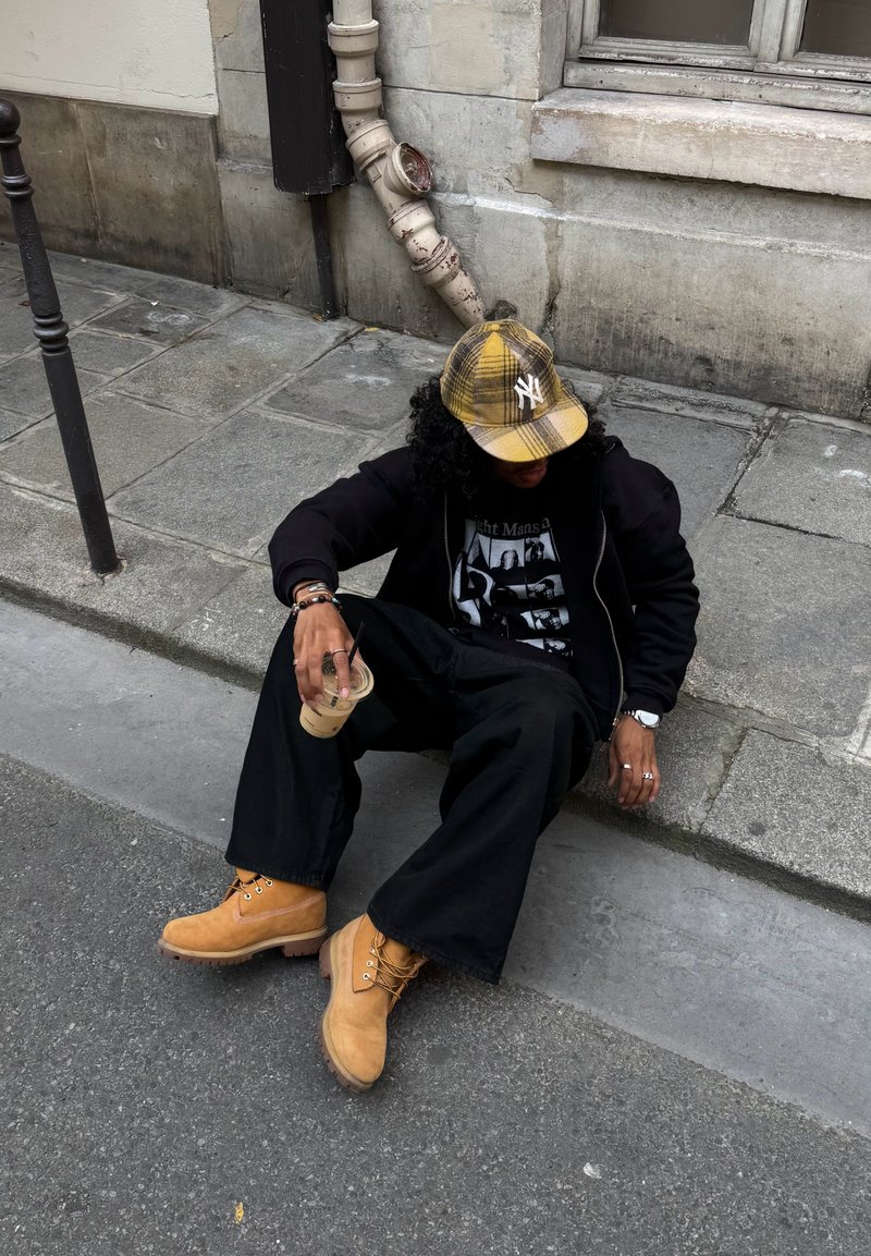 Personne vêtue de vêtements noirs et de bottes couleur beige assise sur un trottoir près du mur d'un bâtiment, tenant un gobelet en plastique avec une paille.
