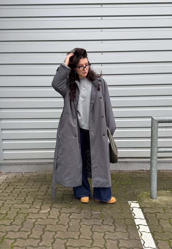 Palton lung gri, cu nasturi, cu rever mari, peste un pulover gri deschis. Blugi evazați de culoare albastră și saboți bej. Stând lângă o balustradă argintie.