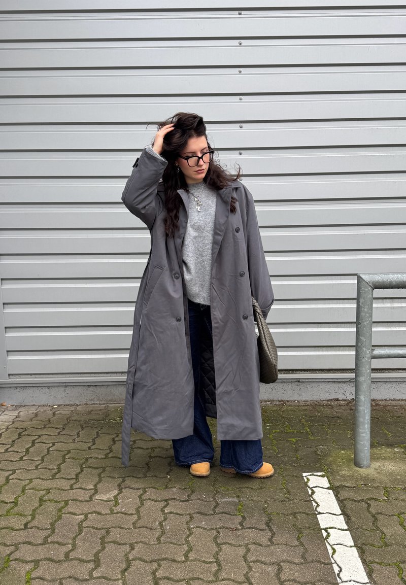Grey long coat, buttoned, with large lapels over a light grey sweater. Flared blue jeans and tan clogs. Standing near a silver railing.