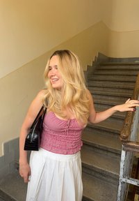 Žena nosi crvenu kariranu bluzu s naborima s bijelom suknjom, drži crnu torbicu, stoji kraj kamenih stepenica s drvenim ogradom.