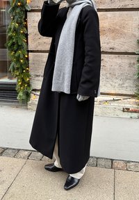 Lang sort uldfrakke med to frontlommer, gråt strikket halstørklæde, cremefarvede bukser og sorte spidse sko. Udendørs miljø med grønt.