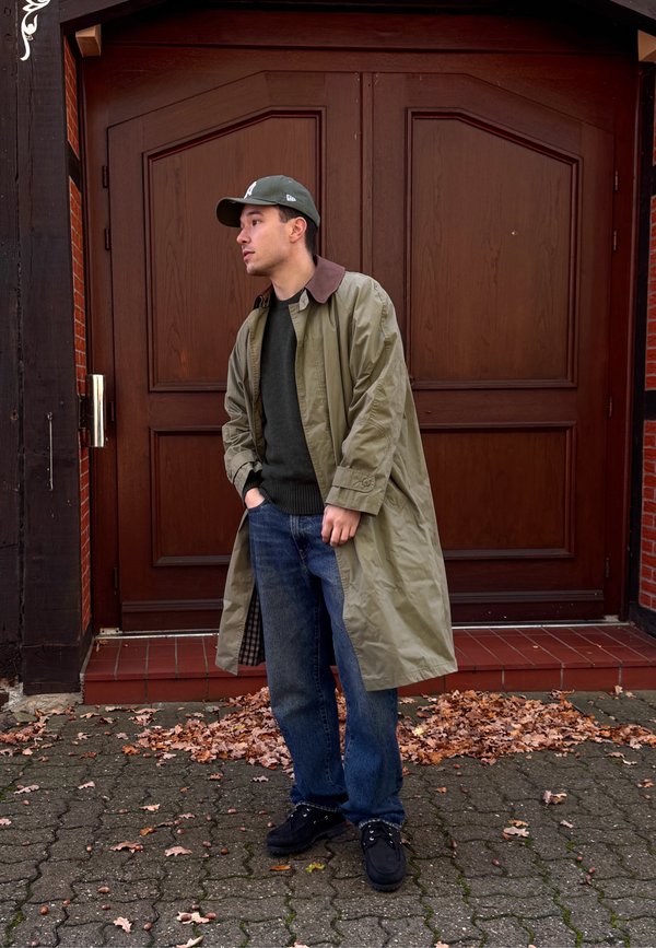 Junger Mann in grünem Trenchcoat, dunklem Pullover, Jeans, schwarzen Schuhen und grünem Hut, der auf einem mit Blättern bedeckten Gehweg vor braunen Holztüren steht.