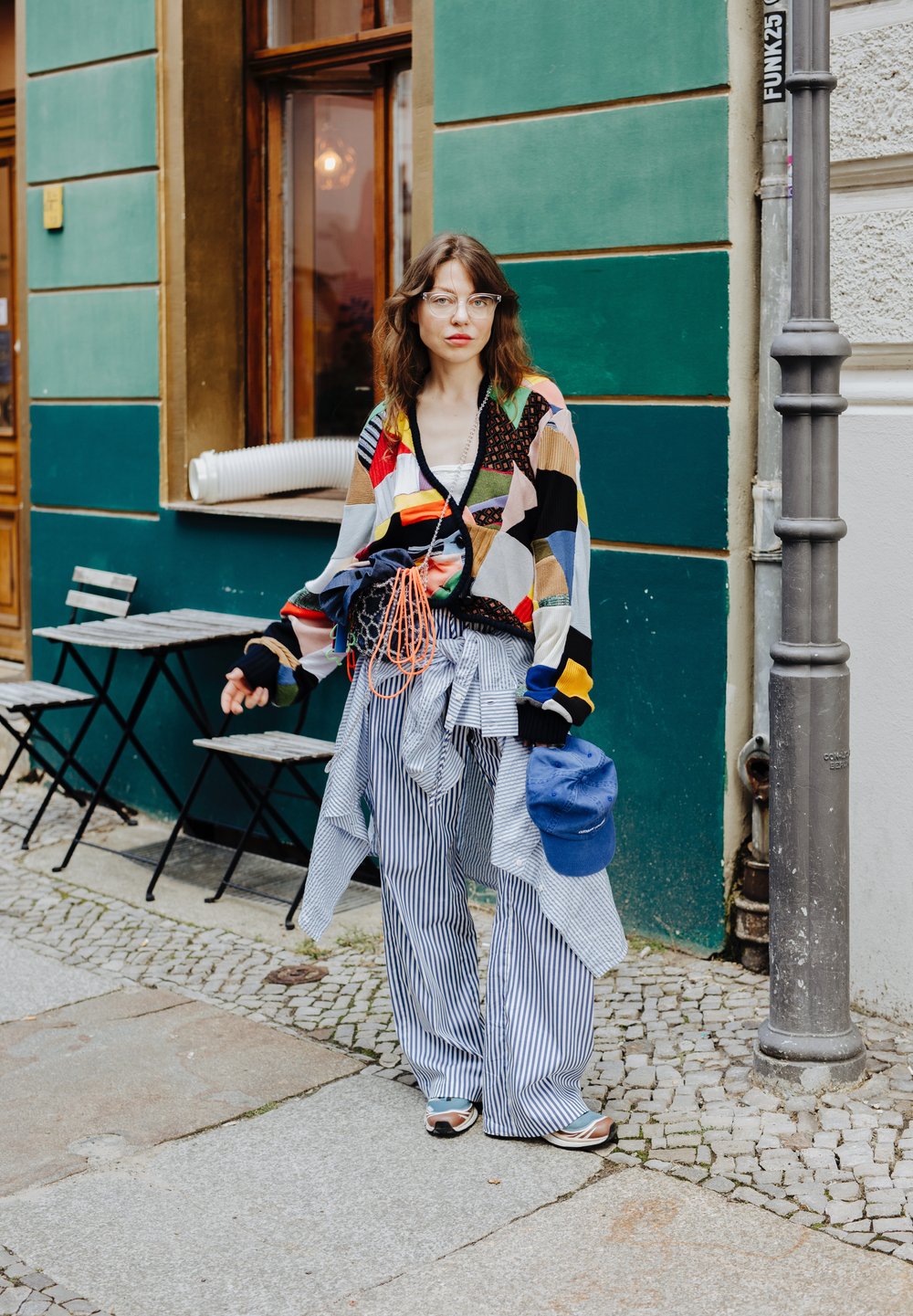 Cardigan oversized com padrão multicolorido, calças listradas em preto e branco, boné azul, acessórios incluem uma bolsa azul e várias peças de roupa.